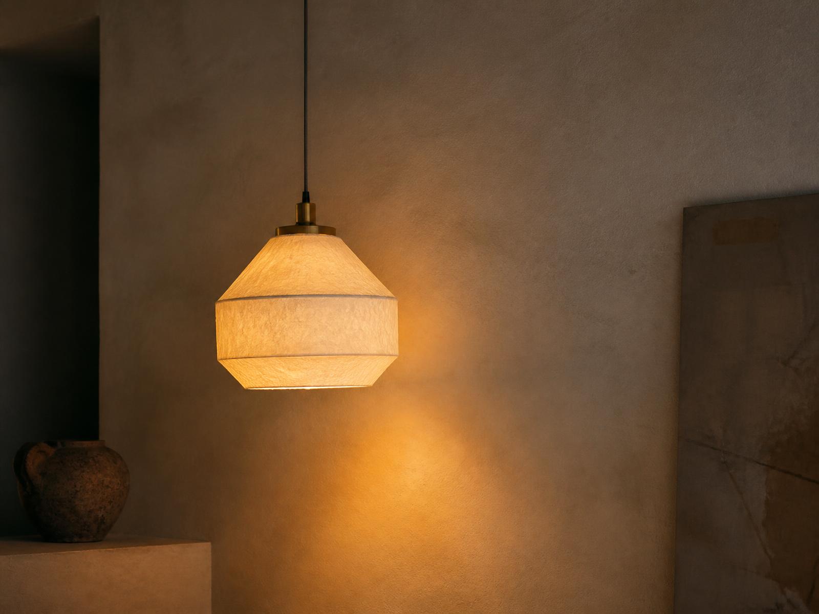 Handmade paper and brass pendant lamp glowing warmly in a dim limewash plaster room