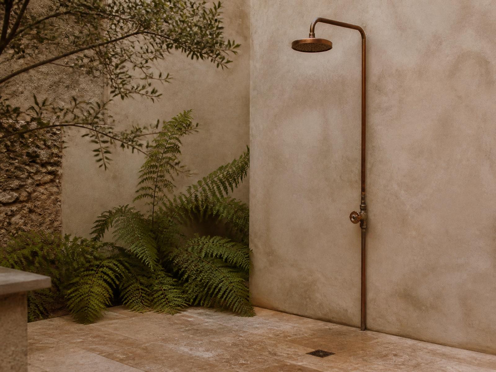 Raw copper outdoor shower mounted on a limewash plaster wall beside ferns on travertine paving